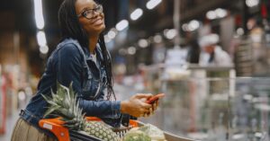 Mulher sorrindo com o celular no supermercado. (Foto: Pexels)