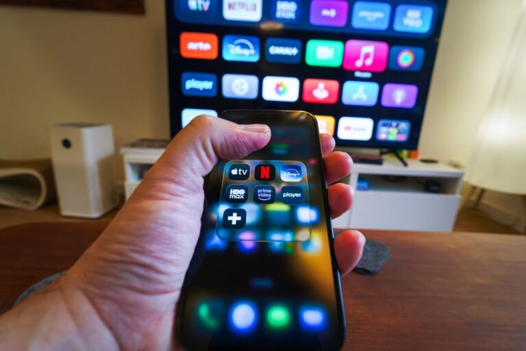 Close-up of a hand holding a phone displaying streaming apps in front of a TV with multiple app icons. Netflix